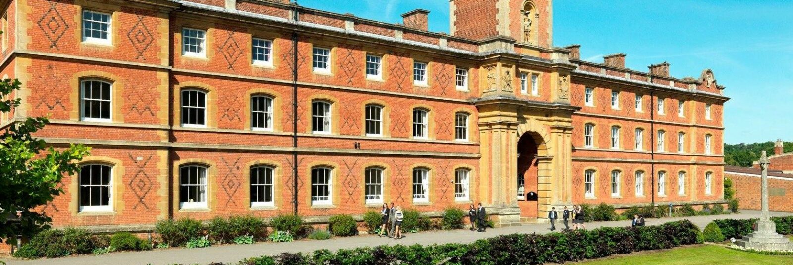 Large historic brick building with three storeys, decorative brickwork, multiple windows, and an arched entrance, with people in front and greenery including bushes and trees, under a clear blue sky.