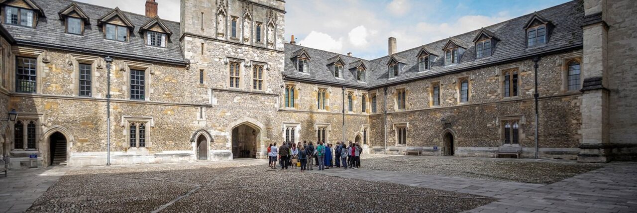 Group of people gathered in the courtyard of a historic stone building with arched doorways, multiple windows, and a slate roof with dormer windows.