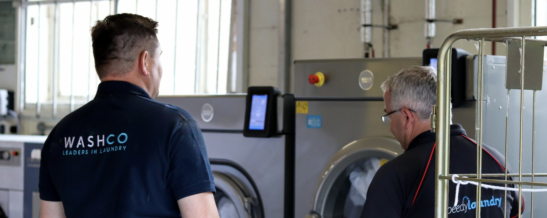 A man in a dark blue WASHCO polo shirt stood next to a man in a speedy laundry top looking at two WASHCONNECT machines