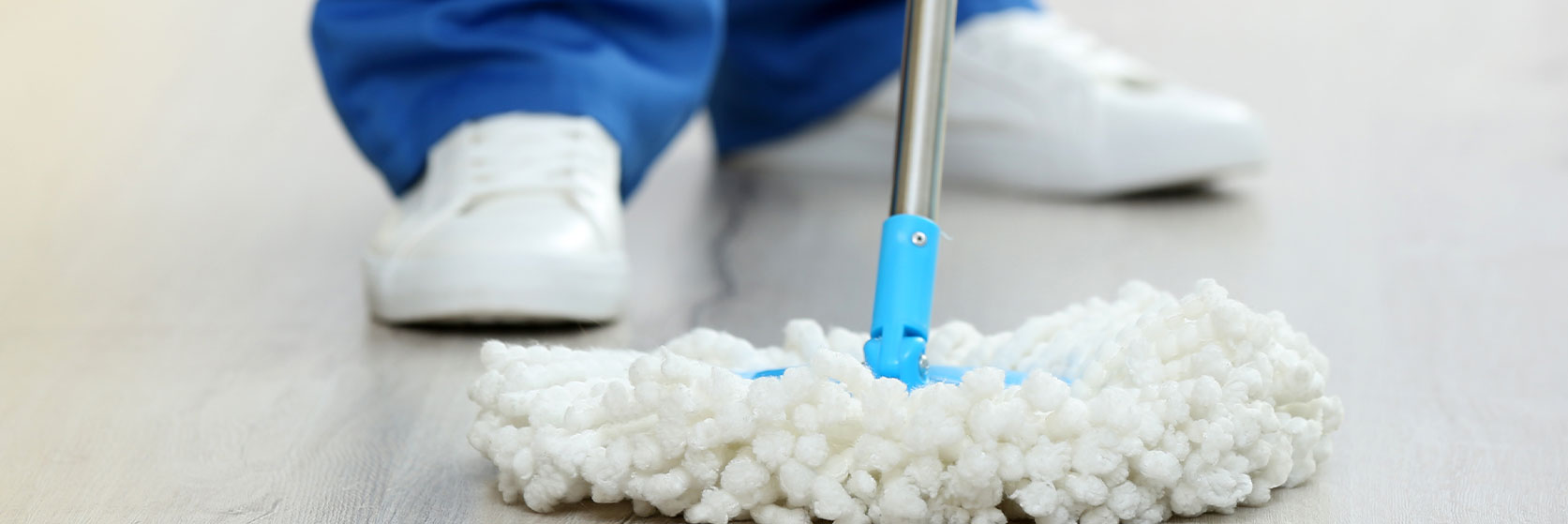 Close-up of a person mopping the floor with a white fluffy mop and blue handle, with their feet visible in white shoes and blue trousers in the background.