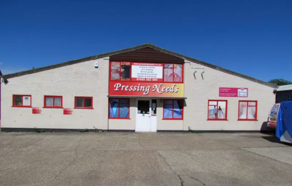 Front of a white building with red trim, displaying a sign for 'Pressing Needs' dry cleaning and laundry services, including service washes, ironing, alterations, and repairs, with contact details and a smaller sign reading 'Board Booth' on the right.
