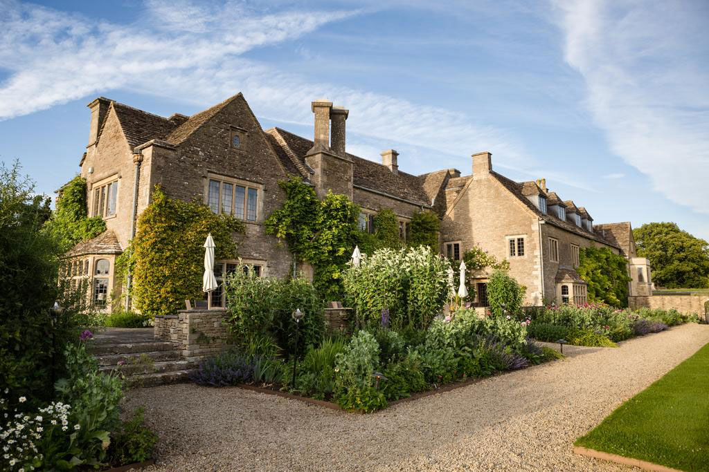 Traditional stone house with multiple chimneys and gabled roofs, surrounded by lush greenery and a well-maintained garden with flowers, shrubs, and white umbrellas for outdoor seating, accessed via a gravel pathway under a clear sky.
