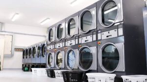 A modern, well-lit laundry room featuring two rows of large, front-loading commercial washing machines and dryers. White and black laundry baskets are lined up on the floor in front of the machines, some of which contain clothing.