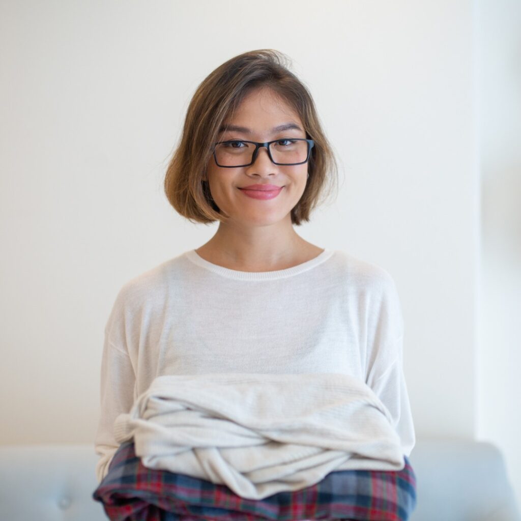 A woman wearing a white shirt and glasses and holding folded clothes, set against a plain light-coloured background.