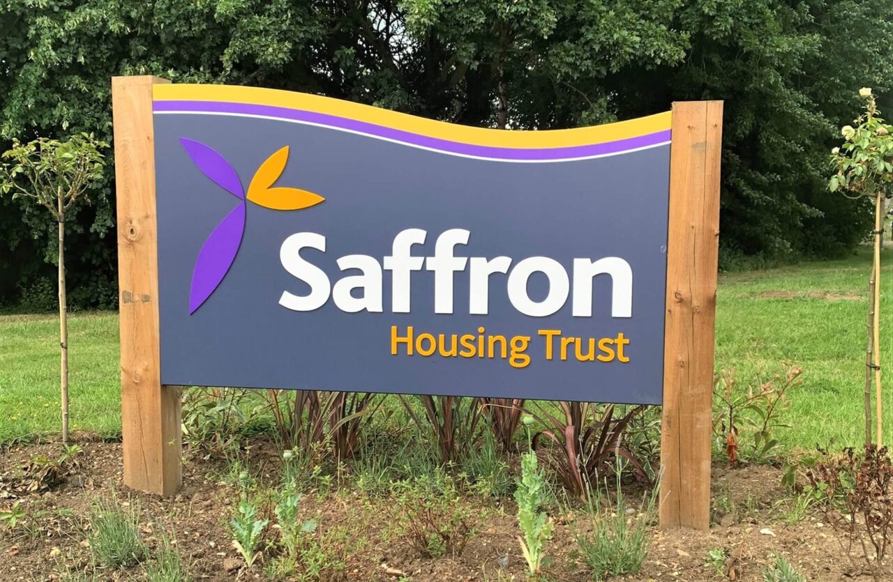 A sign for Saffron housing trust mounted on two wooden posts and stands in a grassy area with plants at the base.