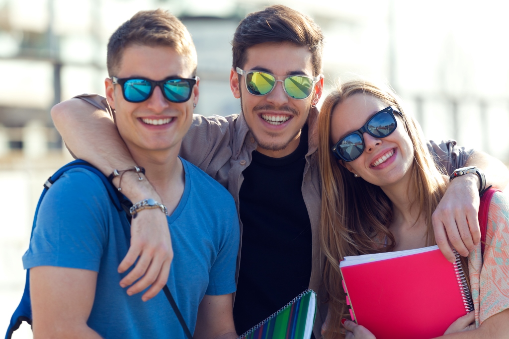 Three people standing close together; the person on the left wears a blue shirt and a backpack, the middle person in black has arms around the others, and the person on the right holds pink and white notebooks or folders.