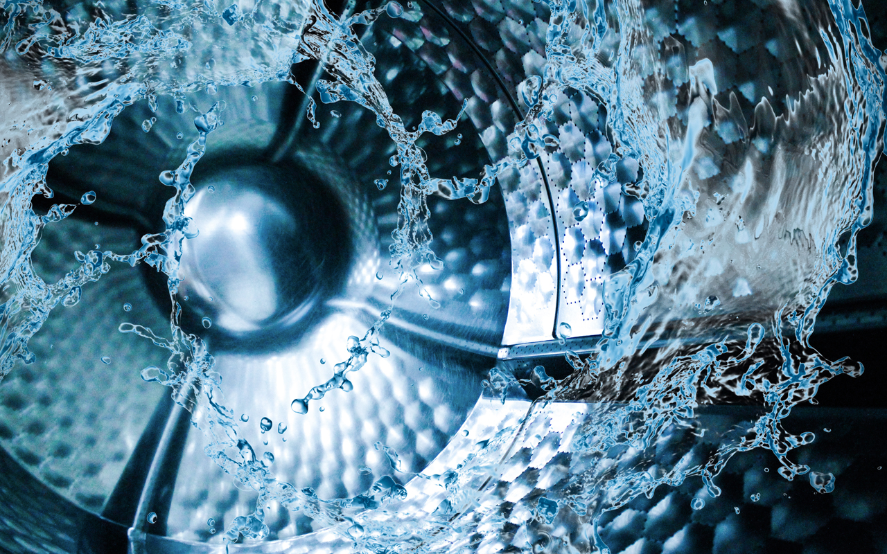 Interior of a washing machine drum with water splashing around, showing a metallic, textured surface and dynamic motion of droplets.