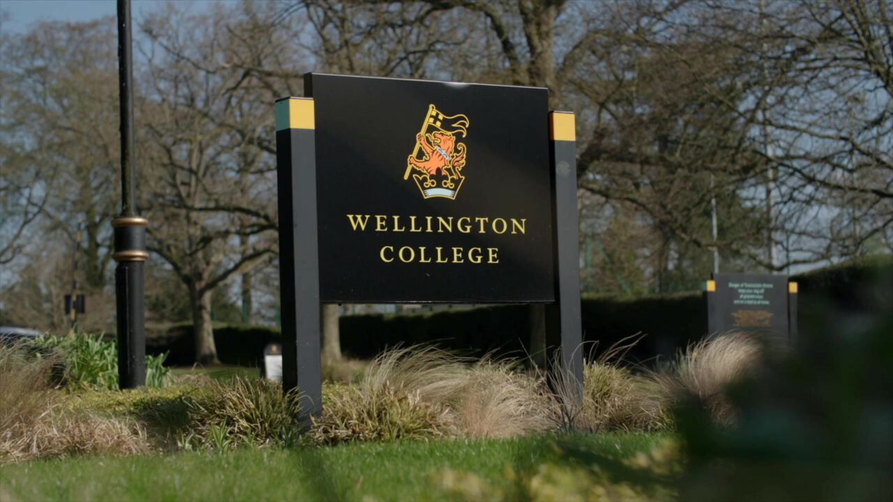 A black sign standing in grass that reads "Wellington College" in yellow writing the the Wellington College logo above it. 