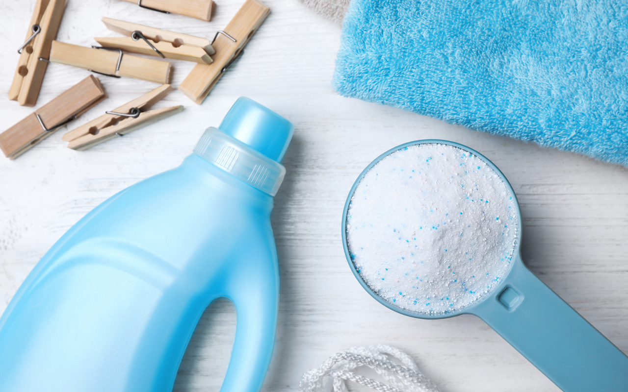Collection of laundry items on a white wooden surface, including a blue bottle of liquid detergent, a scoop of powdered detergent, wooden clothespins, and a folded blue towel.