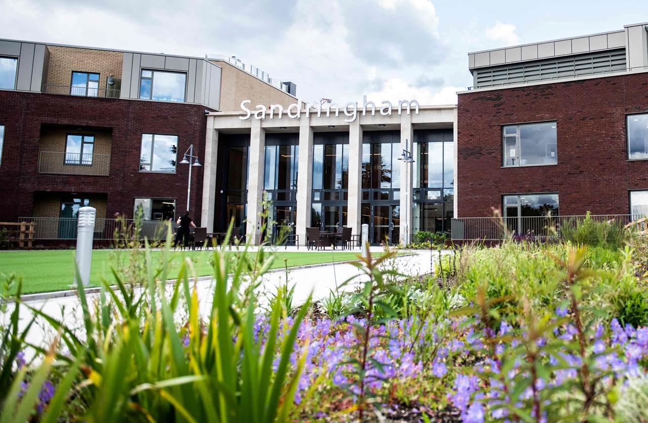 Modern building with a sign reading 'Sandringham' above the entrance, featuring large windows, landscaped gardens with plants and flowers, and visible pathways and outdoor seating areas.