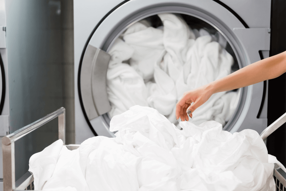 A person loading or unloading white laundry from a front-loading washing machine. The machine is filled with white clothes, and a pile of white laundry is visible in the foreground, possibly in a laundry basket. Only the person's arm is visible, reaching towards the clothes.