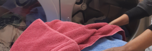 Person handling laundry in front of a washing machine, holding a red towel and a blue towel, with various clothes visible inside the machine.