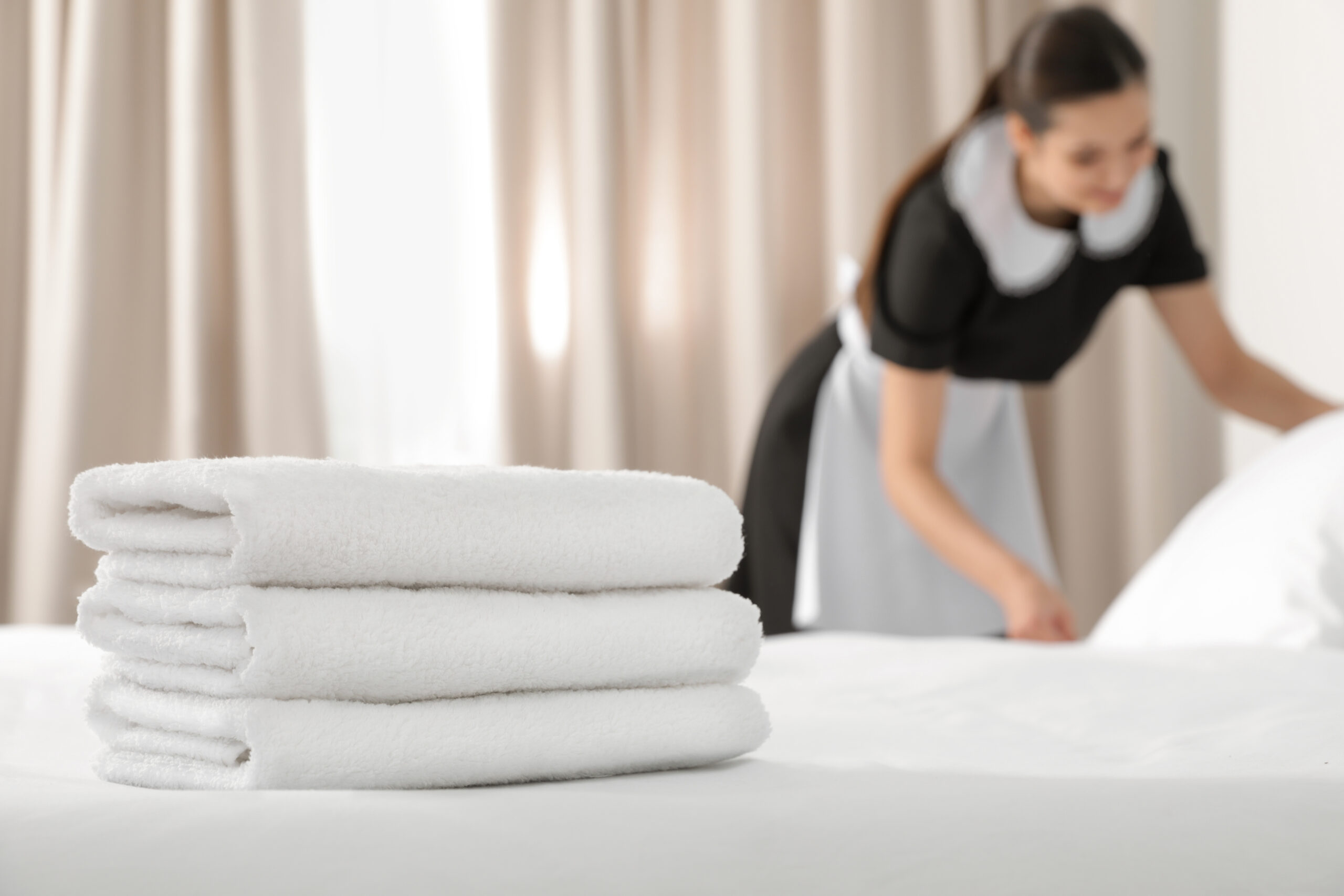 Stacked white clean towels on a hotel bed. A hotel housekeeper can be seen blurred in the background making the bed.