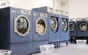 Row of industrial Miele Professional washing machines and dryers in a commercial laundry room, with white laundry baskets placed in front of them.